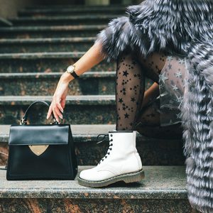 close up legs in white boots of fashionable woman posing in city in warm fur coat with black leather bag